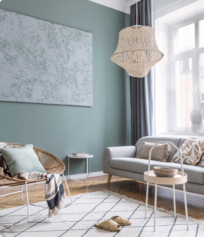 Déco Salon Bohème Moderne : Tissus et Rotin Salon moderne avec mur vert sauge, suspension en rotin, fauteuil en osier et canapé gris clair. Décor bohème et apaisant.