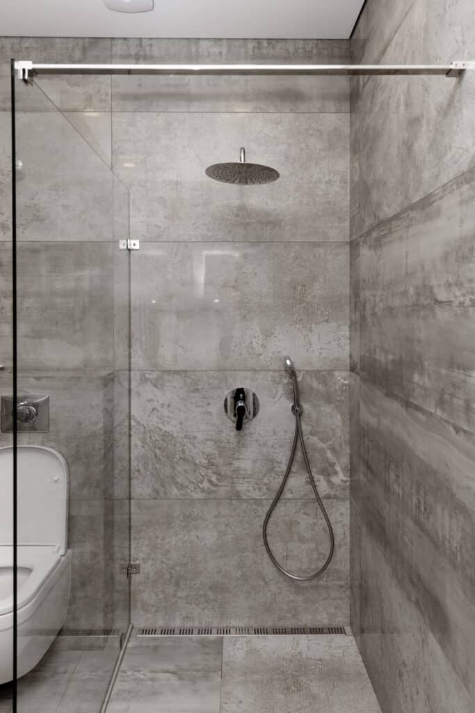 Douche contemporaine carrelage béton et WC suspendu Douche moderne avec parois en verre, carrelage imitation béton gris, pommeau pluie et WC suspendu visible à gauche.