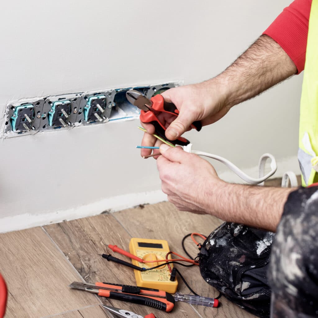 Installation Électrique : Pose de Prises Murales Électricien avec pinces coupantes raccordant des fils électriques colorés dans des boîtiers de prises murales, multimètre au sol.