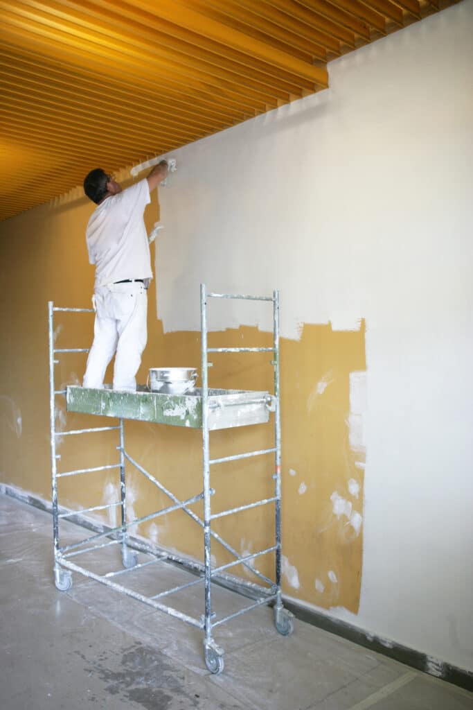 Peintre rénove mur ocre avec sous-couche blanche Peintre en blanc repeint un mur ocre d'une sous-couche blanche depuis un échafaudage roulant sous un plafond jaune.