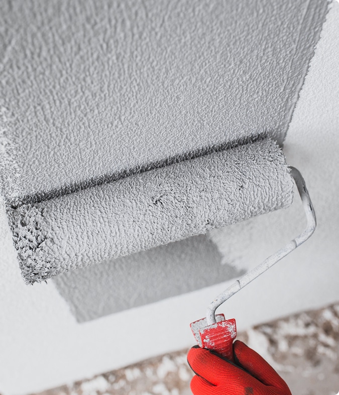 Application de peinture texturée grise au rouleau Application de peinture texturée grise sur un mur ou plafond blanc à l'aide d'un rouleau à poils longs tenu par une main gantée de rouge.