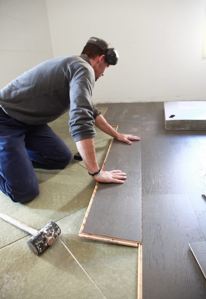 Installation Plancher Flottant Gris : Pose Bricolage Ouvrier agenouillé installant un plancher flottant gris foncé sur une sous-couche verte, un maillet repose à côté.